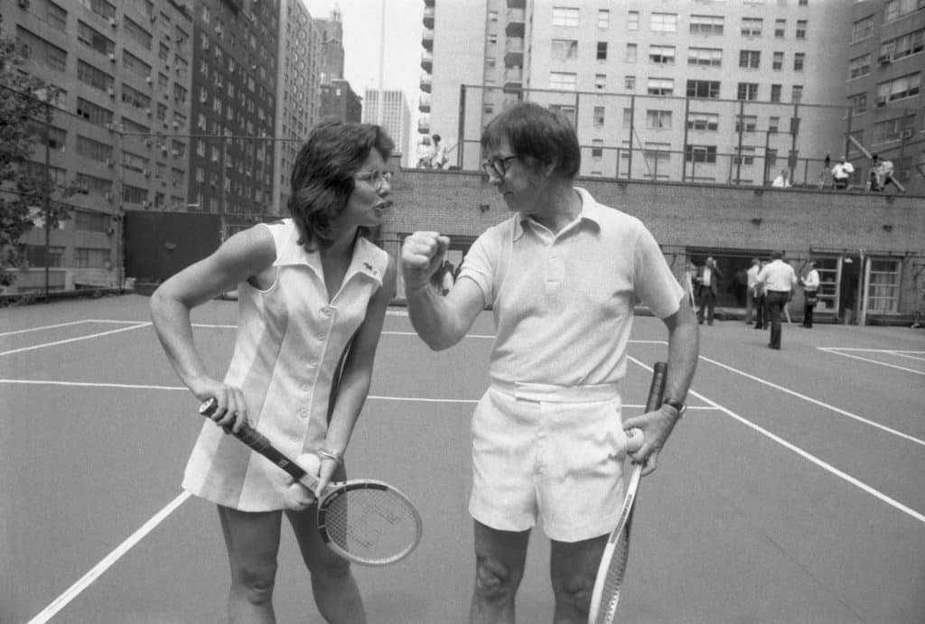 Wimbledon champion Billie Jean King tactfully holds down the net, so that 55-year-old Bobby Riggs can easily clear it during meeting at an east side tennis club here July 11th. Returning in triumph from London, Mrs. King will meet Riggs in a $100,000 winner-take-all tennis match, it was announced July 11th.